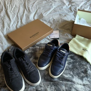 Burberry sneakers i mocka och skinn - Ett par Burberry ett par vagabond sneakers säljes! Ena paret är mörkblått skinn med klassiskt rutigt tyg vid hälen och vita sulor, det andra paret är helsvart mocka med vita sulor. Båda har snörning och låg profil, perfekta för en clean och stilren look.