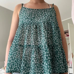 Grön blommig linnetopp med volanger - Supersöt grön linnetopp med vita småblommor och tunna axelband. Toppen är luftig och har volangiga lager som ger en luftig känsla. Perfekt för varma dagar. Säljer pga att den är lite stor.