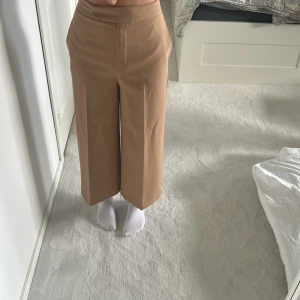 Beiga croppade kostymbyxor från Zara - Snygga beiga kostymbyxor från Zara i croppad modell med hög midja och vida ben. Byxorna har pressveck framtill, diskreta bakfickor och stängs med dragkedja och hake. Perfekta för en clean och trendig look.