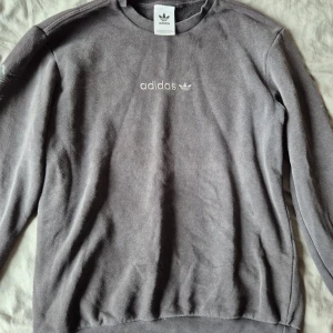 Grå Adidas sweatshirt - Säljer en grå sweatshirt från Adidas med broderad logga på bröstet och tryckta trefoil-loggor längs ärmarna. Tröjan har rund hals, ribbade muddar och är tillverkad i mjuk bomullsmix. Perfekt för en chill och sportig stil.
