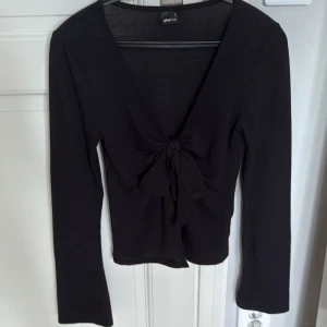 Svart omlottopp med knyt från Gina Tricot - Svart långärmad omlottopp från Gina Tricot med djup v-ringning och knyt framtill. Toppens material är mjukt och stretchigt, vilket ger en slim och smickrande passform. Perfekt inför höst, vinter och fest.🤗
