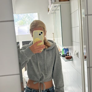 Grå croppad hoodie från NA-KD - Grå croppad hoodie från NA-KD med snörning i midjan och huva. Tröjan har långa ärmar och en relaxed fit som ger en chill vibe. Perfekt att styla med jeans eller kjol för en casual look.