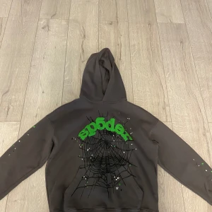 Sp5der hoodie med spindelnätsprint - Mörkgrå hoodie från Sp5der med stor grön logga och svar spindelnät. Hoodien har huva, lång ärm och coola stjärndetaljer på ärmarna. Tillverkad i 100% bomull och har en avslappnad passform. Perfekt för dig som gillar streetwear vibes. Pris kan diskuteras!
