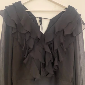 Svart volangblus från Vero Moda - Supersnygg svart blus från Vero Moda med flera lager volanger framtill och v-ringning med knyt i nacken. Den är i storlek L men passar någon som har storlek S 