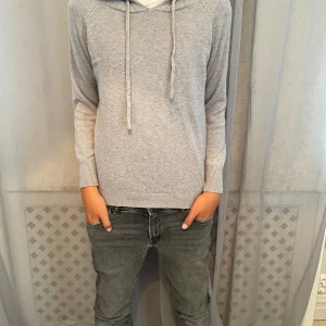 GRÅ MERINOULLS TRÖJA - En stilren ljusgrå merinoulls hoodie med huva och snörning. Tröjan har långa ärmar, ribbade muddar och en mjuk känsla. Perfekt för dig som gillar en enkel och clean look med lite extra mysfaktor.