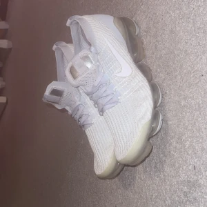 Nike Air Vapormax Flyknit vita sneakers - Nike Air Vapormax Flyknit sneakers i helvitt med stickad ovandel och genomskinlig sula. Skorna har snörning, rund tå och den ikoniska swoosh-loggan på sidan. Superlätta och flexibla med modern design och synlig dämpning i sulan. Kom gärna med bud eller förslag
