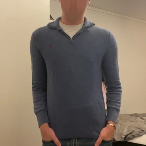Blå stickad tröja Polo Ralph Lauren - Snygg blå stickad tröja från Polo Ralph Lauren med halv dragkedja och klassisk röd broderad logga på bröstet. Tröjan har lång ärm och en stilren krage som ger en clean look. Perfekt för dig som gillar preppy stil och vill ha något bekvämt men ändå dressat. Passar storlek XS!