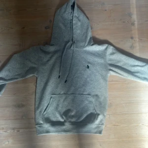 Grå hoodie från Polo Ralph Lauren - Snygg grå hoodie från Polo Ralph Lauren med klassisk svart broderad logga på bröstet. Tröjan har huva med snörning, stor magficka och är tillverkad i mjuk bomull. Perfekt för en chill och stilren look. Passar om du är 155-165