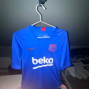 Blå FC Barcelona Nike fotbollströja - Säljer en blå FC Barcelona fotbollströja från Nike med klubbmärke och röd Nike-logga på bröstet. Tröjan har korta ärmar, vit BEKO-sponsor på framsidan och en röd detalj i nacken. Tillverkad i lätt och ventilerande funktionsmaterial.