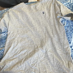 Grå t-shirt från Polo Ralph Lauren - Snygg grå t-shirt från Polo Ralph Lauren med klassisk blå logga broderad på bröstet. T-shirten har rund halsringning och korta ärmar, perfekt för en clean och enkel stil. Tillverkad i mjuk bomull som känns skön mot huden.