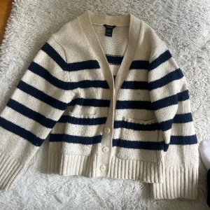 Randig beige kofta från Lindex XS - Supermysig stickad kofta från Lindex i beige med mörkblå breda ränder. Koftan har v-ringning, knappar framtill och två fickor. Perfekt för lager-på-lager och chill dagar. Tjock ribbad mudd vid ärmslut och nederkant.