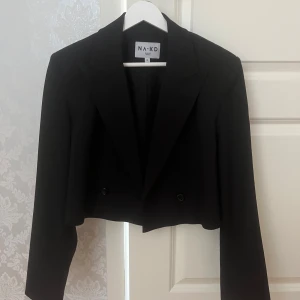 Svart croppad kavaj från NA-KD - Snygg svart croppad kavaj från NA-KD med klassisk krage och dubbelknäppning. Jackan har lång ärm och en stilren, rak passform som ger en modern look. Perfekt att styla med både kjol och byxor 💓💓 i ny skick💓