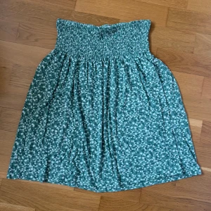 Grön blommig kortkjol med smock - Supersöt grön kortkjol med vitt blommönster och bred smock i midjan. Kjolen är luftig och har en flowy passform som är perfekt för varma dagar. Passar dig som gillar en avslappnad men ändå söt stil. Från Indiska