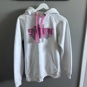 Vit hoodie med Sälen tryck - Vit hoodie med rosa detaljer och broderad text 'Sälen' framtill. Stora rosa snören i huvan och en praktisk magficka. Mjuk insida och klassisk passform, perfekt för chill dagar. Snygg logga på huvan och coolt tryck i silver och rosa.