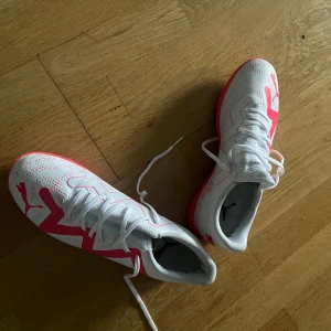 Vita och röda fotbollsskor från Puma - Snygga vita fotbollsskor från Puma med rosa detaljer och logga på sidan. Skorna har snörning och en platt sula som passar perfekt för inomhusspel. Ovandelen är i syntetmaterial med perforeringar för extra ventilation. Inga defekter och är som ny! Använd fåtal gånger! (Futsalskor)