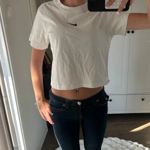 Vit croppad t-shirt från Nike - Säljer en vit croppad t-shirt från Nike med svart broderad swoosh på bröstet. T-shirten har korta ärmar, rund halsringning och är gjord i mjuk bomull. Perfekt till jeans eller shorts för en chill look.