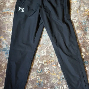 Svarta träningsbyxor Under Armour - Svarta träningsbyxor från Under Armour med logga på vänster lår. Byxorna har en smal passform och är gjorda i ett lätt och funktionellt material som passar perfekt för träning eller chill. Elastisk midja och diskreta fickor på sidorna.