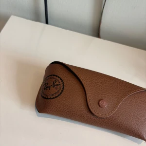 Ray-Ban svarta solglasögon med fodral (helt nya) - Snygga svarta Ray-Ban solglasögon med klassisk rundad form. Kommer med original brunt fodral i läderlook och putsduk. Perfekta för att lyfta din stil och skydda ögonen mot solen. Ikonisk logga på glaset och stilren design.