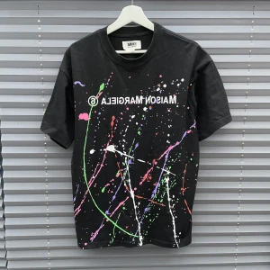 Maison Margiela T-shirt - Svart t-shirt från Maison Margiela med coola färgstänk i grönt, rosa, vitt, lila och rött. Trycktet av Maison margiela på bröstet och klassisk passform. Storlek: 44 (M/L). Skulle behöva tvättas en gång men annars helt felfri. Bara komma dm vid frågor och funderingar!!!