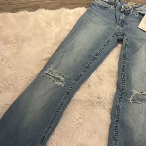 Ljusblå jeans från Nelly med slitningar - Ljusblå jeans från Nelly med coola slitningar på benen och snygga broderade detaljer på bakfickorna. Klassisk femficksmodell med normal passform och dragkedja. Perfekta för en avslappnad och trendig look.