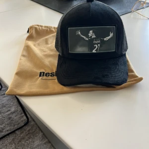 Zlatan trycker keps  - Svart keps med mesh på sidorna och böjd skärm. Framtill finns ett stort svartvitt fotomotiv av en fotbollsspelare med utsträckta armar och nummer 27 på ryggen. Kepsen har justerbar baksida och levereras med en gul tygpåse från Designhallen.se. Aldrig använd !