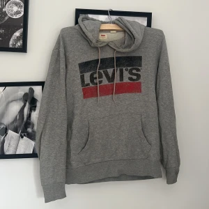 Grå hoodie från Levi's med logga - Säljer en grå hoodie från Levi's i storlek S. Tröjan har klassisk logga framtill i svart och rött, huva med snörning och en stor magficka. Perfekt för chill dagar och enkel att matcha med jeans eller joggers.