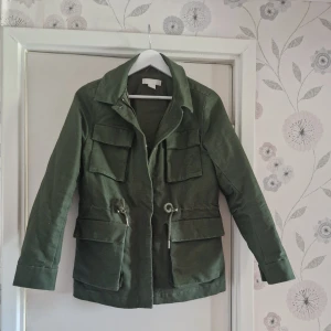 HM Cotton cargo jacket - Snygg grön cargo jacka  med fyra stora fickor framtill och justerbar snörning i midjan. Jackan har klassisk krage och långa ärmar, perfekt för lager-på-lager-stil. Passar dig som gillar en avslappnad men ändå stilren look.