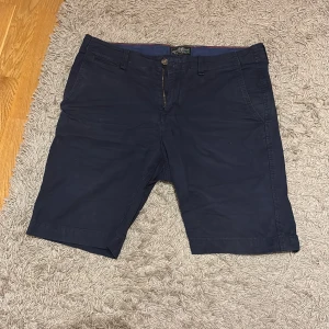 Marinblå shorts från Hampton Republic - Snygga marinblå shorts från Hampton Republic med klassisk passform. De har fickor fram och bak, bälteshällor och stängs med knapp och dragkedja. Perfekta för varma dagar när du vill ha en stilren look.