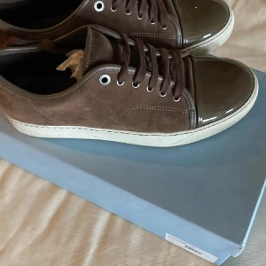 Bruna lanvin - Snygga bruna sneakers med ovandel i mocka och en glansig, lackad tådel. Skorna har vita sulor och bruna skosnören. Perfekt för dig som gillar en stilren men ändå unik look. 