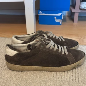 Bruna Saint Laurent sneakers i mocka - Snygga bruna sneakers från Saint Laurent i mocka med vita detaljer på hälen och vita snören. Klassisk låg modell med rund tå och platt sula. Perfekta för dig som gillar stilrena och tidlösa skor med lyxig känsla. Skorna är välanvända därav det låga priset, men fortfarande väldigt mycket kvar och ge! Nypris: Ca 7000
