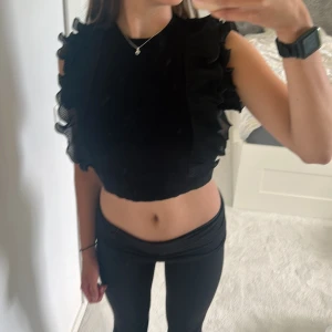 Svart volangtopp från NA-KD - Supersnygg svart topp från NA-KD med breda plisserade volanger längs axlarna. Modellen är ärmlös och har en croppad passform. Materialet är stickat och volangerna är i ett tunt, transparent tyg som ger en cool kontrast.