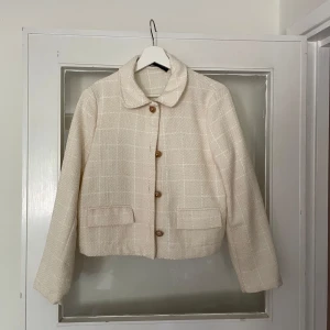 Beige rutig kavaj med knappar - Stilren beige kavaj i rutigt mönster med snygga guldknappar framtill. Jackan har klassisk krage, två fickor och är croppad i modellen. Använd 2-3 ggr. 