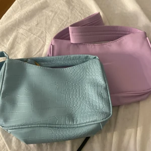Pastellblå och lila axelväskor - Två snygga axelväskor i pastellfärger. Den ena är ljusblå med croco-mönstrad yta och den andra är lila med slät finish. Båda har enkel, stilren design och bred axelrem. Perfekta för att lyfta din outfit med färg och trendig vibe.