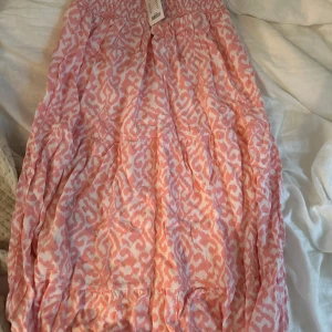 Rosa mönstrad kjol från 157 - Supersöt rosa och vit kjol från 157 med elastisk smock i midjan och ett unikt abstrakt mönster. Kjolen är luftig och har volanger som ger en skön och trendig look. Perfekt för dig som gillar färg och vill sticka ut lite extra.