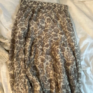 Blommig långkjol från Sofie Schnoor - Supersöt långkjol från Sofie Schnoor med beige botten och brunt blommönster. Kjolen har en avslappnad passform med resår i midjan och är gjord i ett lätt bomullstyg. Perfekt för dig som gillar romantiska och bohemiska vibes.