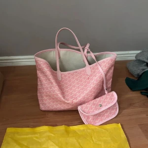 Handväska köpt utomlands - Säljer en stor rosa shoppingväska med klassiskt grafiskt mönster och matchande liten clutch. Väskan har två smala handtag och är tillverkad i syntetmaterial med läderdetaljer. Perfekt för dig som vill sticka ut med en färgglad och ikonisk accessoar. Superfint skick, köpt för 2200