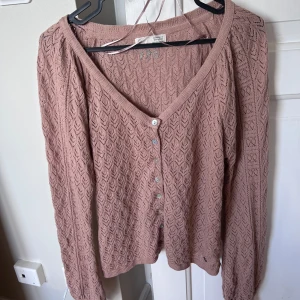 Stickad kofta i puderrosa från Odd Molly - Supersöt puderrosa stickad kofta från Odd Molly med v-ringning och fina knappar framtill. Koftan har ett jättefint hålmönster. Kommer med en extra knapp ifall någon skulle ramla av.