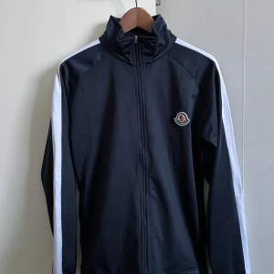 Moncler svart vindjacka med vita ränder - Svart vindjacka från Moncler med vita ränder längs ärmarna och klassisk logga på bröstet. Jackan har hög krage, hel dragkedja och sportig look. Perfekt för dig som gillar stilrena och exklusiva plagg med streetkänsla.