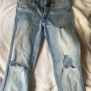 Ljusblå bootcut jeans med hål - Säljer ett par bootcut, ljusblå jeans med slitna detaljer och stora hål på båda benen. Jeansen har dubbla knappar i midjan, dragkedja och klassiska fickor. Bra kvalitet och har använts ett fåtal gånger. St 152, passar uppåt 160.