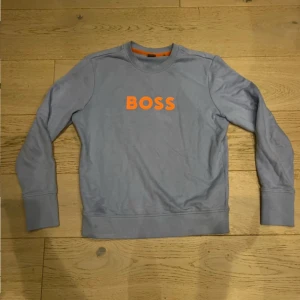 Ljusblå BOSS sweatshirt med orange tryck - Snygg ljusblå sweatshirt från BOSS med orange BOSS-tryck på bröstet. Tröjan har rund halsringning, långa ärmar och breda muddar vid ärmslut och nederkant. Perfekt för en chill och stilren look.