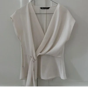 Vit omlottopp från Zara - Säljer en stilren vit omlottopp från Zara med knyt framtill. Toppen är ärmlös och har en snygg v-ringning. Perfekt för dig som gillar en clean och fräsch look. Lätt och luftig i materialet.