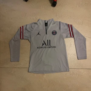 PSG långärmad träningströja Jordan - Snygg långärmad PSG träningströja i ljusgrått med svarta och röda detaljer på ärmarna. Tröjan har halv dragkedja, PSG-logga på bröstet och Jordan-logga. Tillverkad i ett lätt och stretchigt funktionsmaterial. Passar M då den sitter lite tajt och priset in diskuteras.