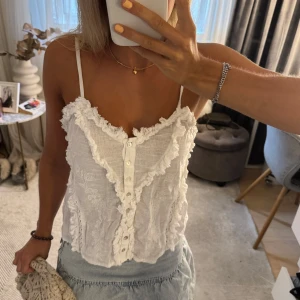 Vit broderad linnetopp med volang - Superfin vit linnetopp med smala axelband och broderade blommor. Toppen har volangdetaljer längs urringningen och framtill, samt knappar i mitten. Lätt och luftig med romantisk känsla, perfekt för varma dagar.