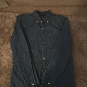 Jeansskjorta från Tommy Hilfiger - Snygg mörkblå jeansskjorta från Tommy Hilfiger med klassisk krage och knappar framtill. Liten broderad logga på bröstet och långa ärmar. Perfekt för dig som gillar en stilren och tidlös look. Passar perfekt för dig som är mellan 148-154cm. Perfekt för sommarkvällar🌆