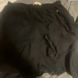 Svart volangkjol med resårmidja - Supersöt svart kortkjol med volangdetaljer och resår i midjan. Kjolen har en flowy känsla och passar perfekt till en trendig look. Enkel att matcha med både sneakers och boots för en cool vibe.