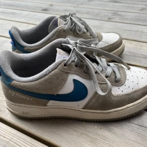 Nike Air Force 1 grå/vit/ blå - Nike Air Force 1 sneakers i grå mocka och vitt skinn med blå swoosh på sidorna. Klassisk låg modell med grå snörning och perforerad tå för extra ventilation. Snygg retrostil som passar perfekt till streetwear och casual outfits.