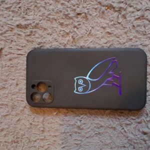 Svart OVO mobilskal med uggla - Säljer ett svart mobilskal med OVO:s ikoniska uggla i holografisk lila och blå nyans. Skalet är gjort i mjuk plast och har öppningar för kamera och knappar. Perfekt för dig som vill ha en clean look med lite extra stil.