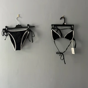 Svart bikini med vita detaljer från NA-KD i storlek XXS💕 - Supersnygg svart bikini från NA-KD med vita virkade kanter. Klassisk trekantsmodell upptill och knyt på både över- och underdel. Perfekt för stranden eller poolen, med trendig kontrast mellan svart tyg och vita detaljer.