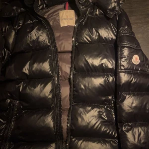 Svart Moncler pufferjacka med huva - Snygg svart pufferjacka från Moncler med glansig finish och klassisk logga på ärmen. Jackan har huva, dragkedja framtill och breda quiltade sektioner. Perfekt för kalla dagar när du vill hålla stilen och värmen. Priset är diskutabelt 