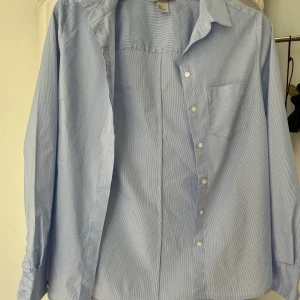 Blåvit randig skjorta från H&M - Klassisk blåvit randig skjorta från H&M i storlek 36. Skjortan har lång ärm, bröstficka och knäppning framtill. Tillverkad i bomull för en skön och luftig känsla. Perfekt till jeans eller kjol för en clean look.